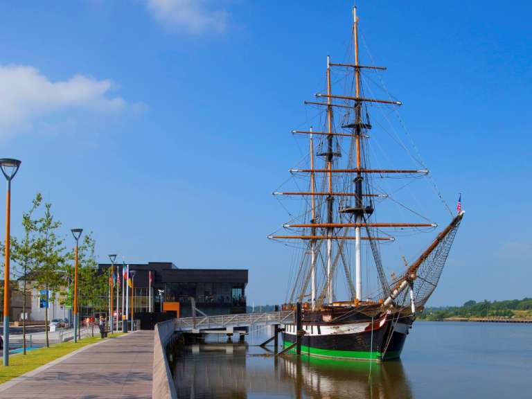 Ein Segelschiff mit vielen Masten liegt vor einem modernen Gebäude an einem sonnigen Tag am Wasser.