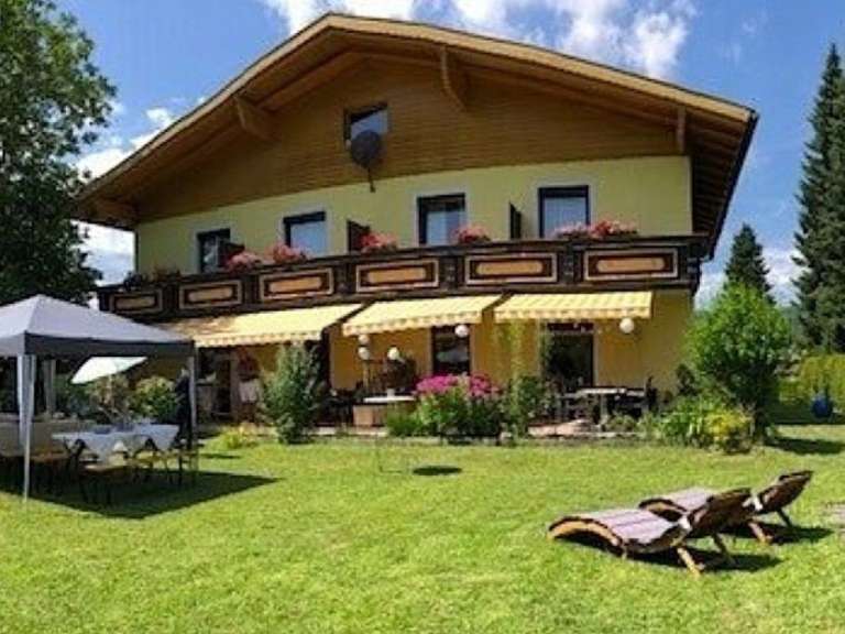 Auf einer grünen Wiese vor einem gelben Haus mit Holzbalkon stehen zwei Liegestühle, während im Vordergrund ein Pavillon mit Tischen und Stühlen zu sehen ist.