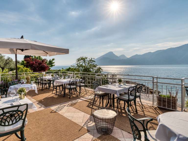 Auf einer Terrasse mit Blick auf das Meer und die Berge sind mehrere Tische für eine Mahlzeit gedeckt.