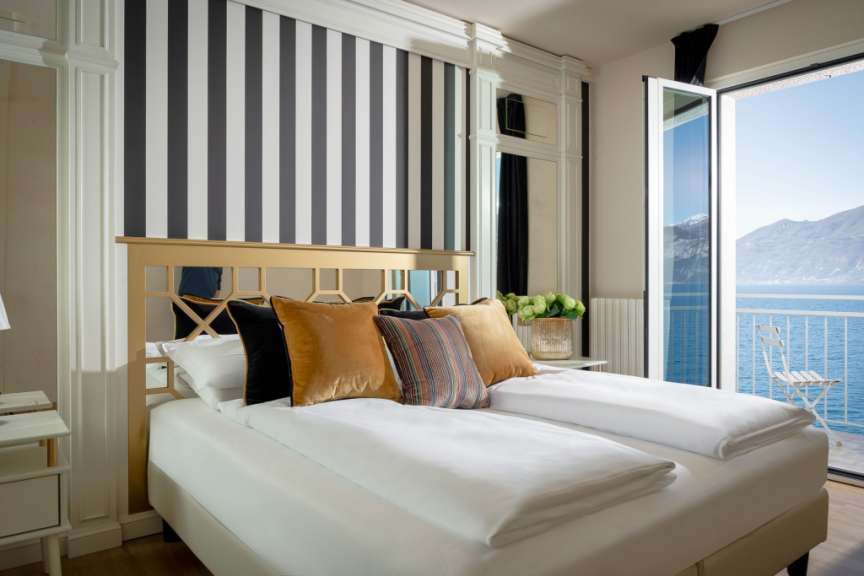 Ein Doppelbett mit weißer Bettwäsche und Kissen steht vor einer gestreiften Wand mit Blick auf das Meer und die Berge.