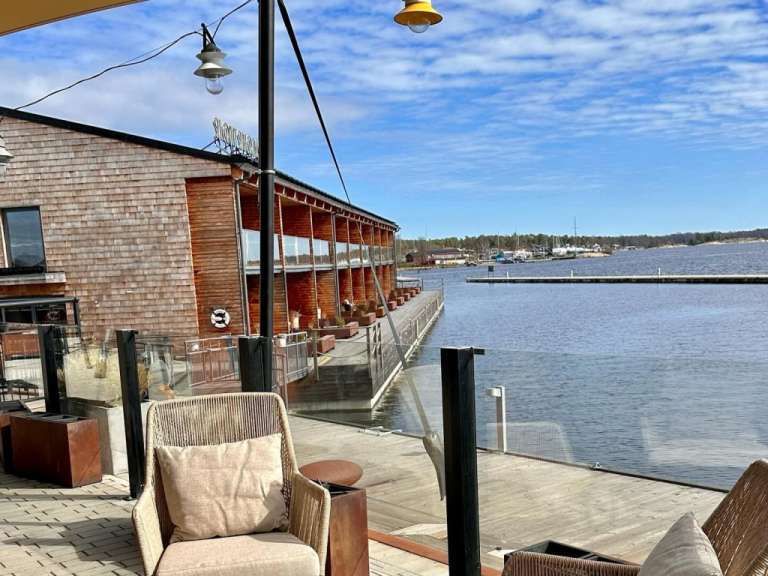 Auf dem Bild ist eine Terrasse mit Blick auf ein Gewässer mit Booten und einer Holzgebäudefassade zu sehen.