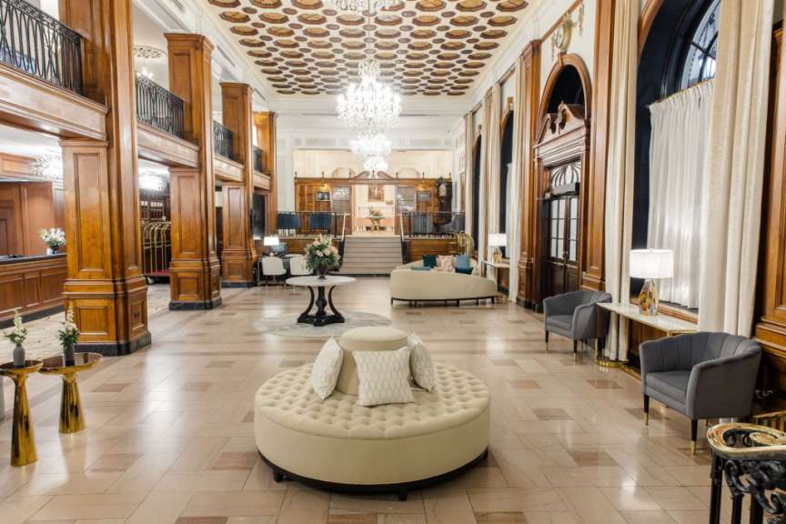 Eine luxuriöse Hotellobby mit einem runden, gepolsterten Sofa, zwei Sesseln, einem Couchtisch und einer eleganten Decke mit Kronleuchtern.