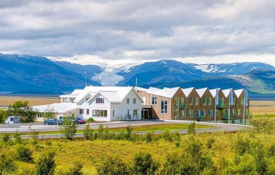 Ein modernes Hotel mit weißem und holzfarbenem Fassadenbau steht in einer grünen Landschaft vor einer Bergkulisse mit Gletschern.