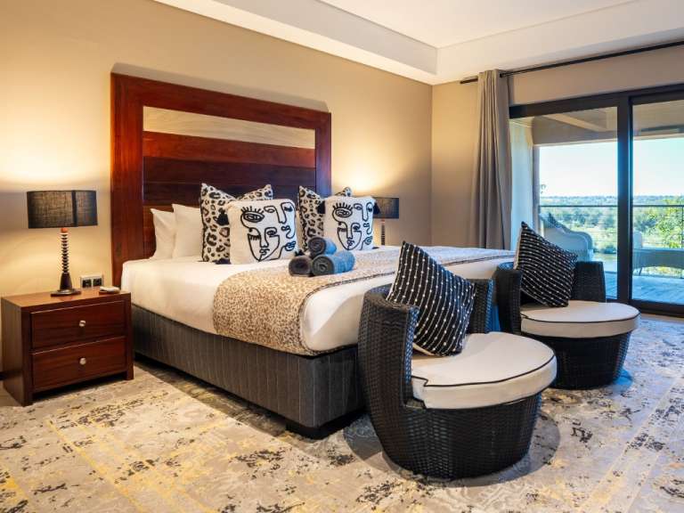 Das Bild zeigt ein luxuriöses Schlafzimmer mit einem großen Bett, zwei Sesseln und einem Blick auf eine offene Landschaft.
