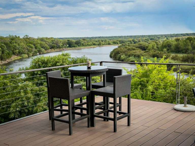 Ein Tisch mit vier Stühlen steht auf einer Holzterrasse mit Blick auf einen Fluss und eine grüne Landschaft.