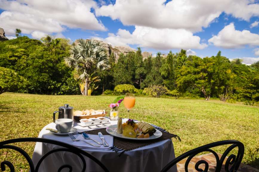 Ein Frühstückstisch mit Früchten, Gebäck und Saft steht auf einer Terrasse mit Blick auf eine üppige grüne Landschaft unter einem bewölkten blauen Himmel.