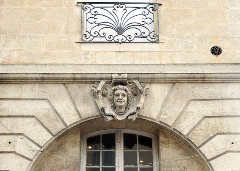 Eine Steinfassade mit einem Fenstergitter, einer geschnitzten weiblichen Büste und einem Bogen über einem Fenster.