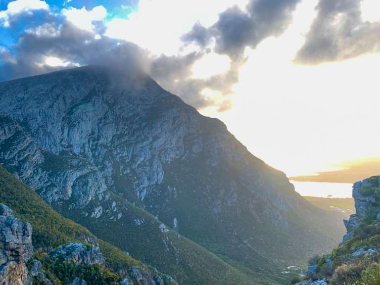 Ein dramatischer Berg mit steilen, felsigen Hängen und bewaldeten Tälern wird vom warmen Licht der tief stehenden Sonne beleuchtet.