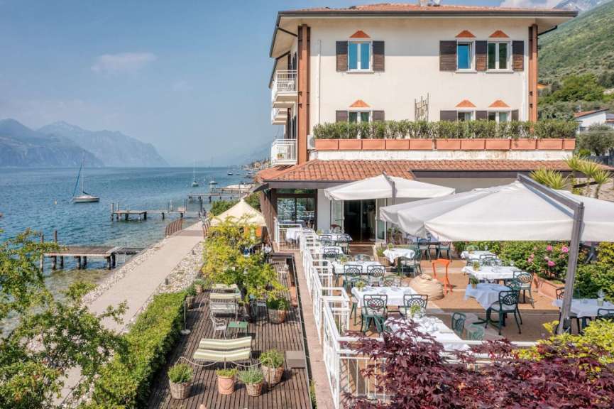 Ein Restaurant mit Blick auf einen See und Berge, im Vordergrund Tische und Stühle im Freien.