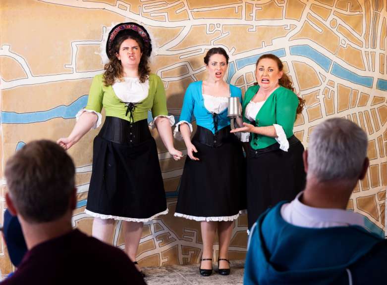 Drei Schauspielerinnen stehen vor einer Bühnenwand, die eine stilisierte Stadtkarte darstellt, und schauen mit expressionistischen Grimassen ins Publikum.