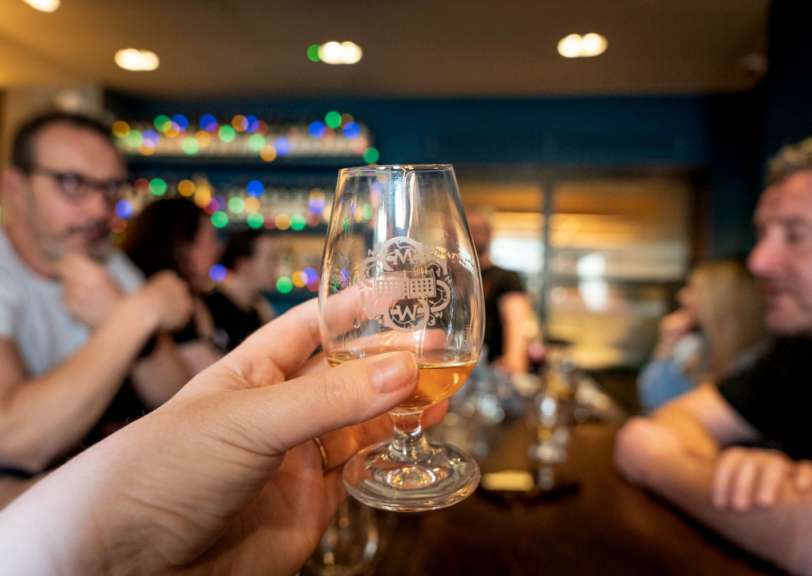 Eine Hand hält ein kleines Glas mit einem bernsteinfarbenen Getränk vor einer verschwommenen Kulisse von Menschen und bunten Lichtern.