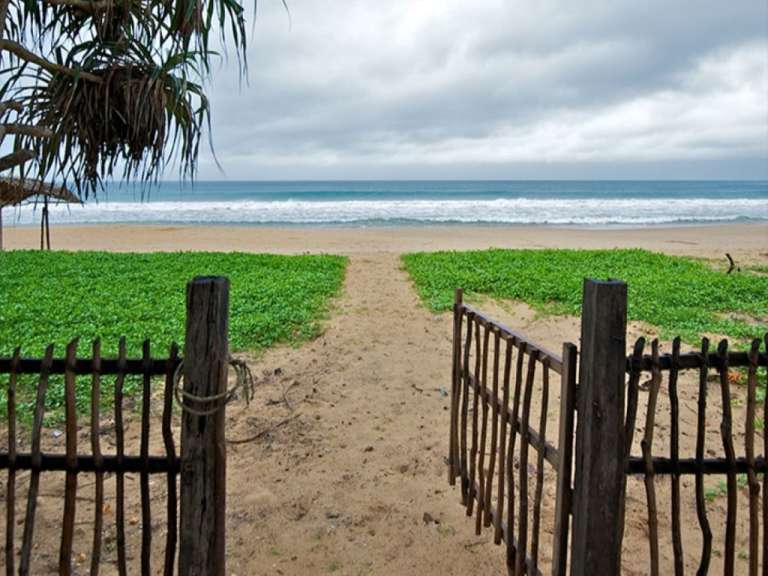 Ein sandiger Weg führt durch einen hölzernen Zaun und üppiges grünes Gestrüpp zu einem ruhigen Strand mit azurblauem Meer unter einem bedeckten Himmel.