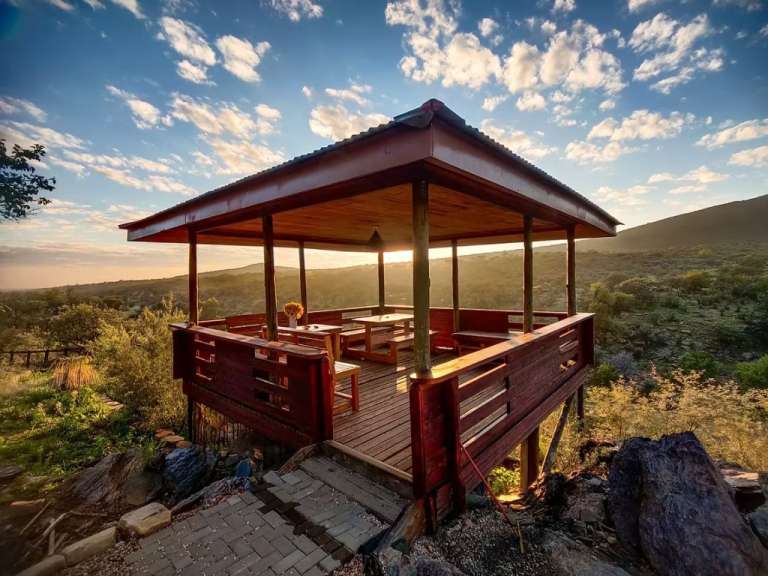 Eine hölzerne Veranda mit Sitzgelegenheiten und Tischen steht auf einem felsigen Hügel mit Blick auf eine hügelige Landschaft und einen bewölkten Himmel bei Sonnenuntergang.