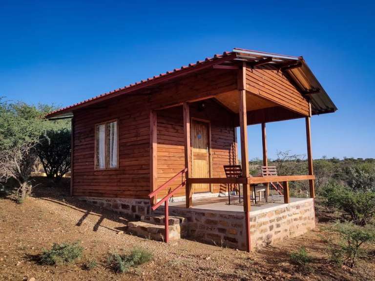 Ein kleines Holzhaus mit Veranda und zwei Stühlen steht auf einem Hügel unter einem blauen Himmel.