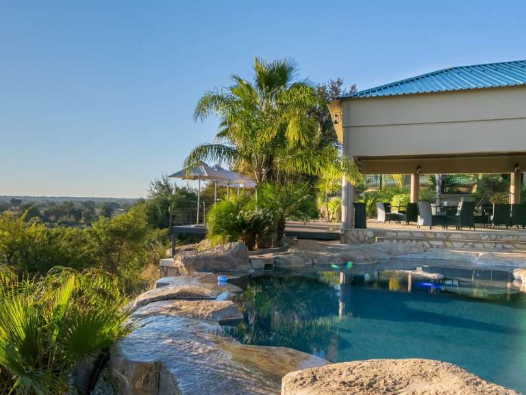 Ein türkisfarbener Pool umgeben von Steinen und tropischer Vegetation mit einer überdachten Terrasse und einem Essbereich im Hintergrund unter einem blauen Himmel.