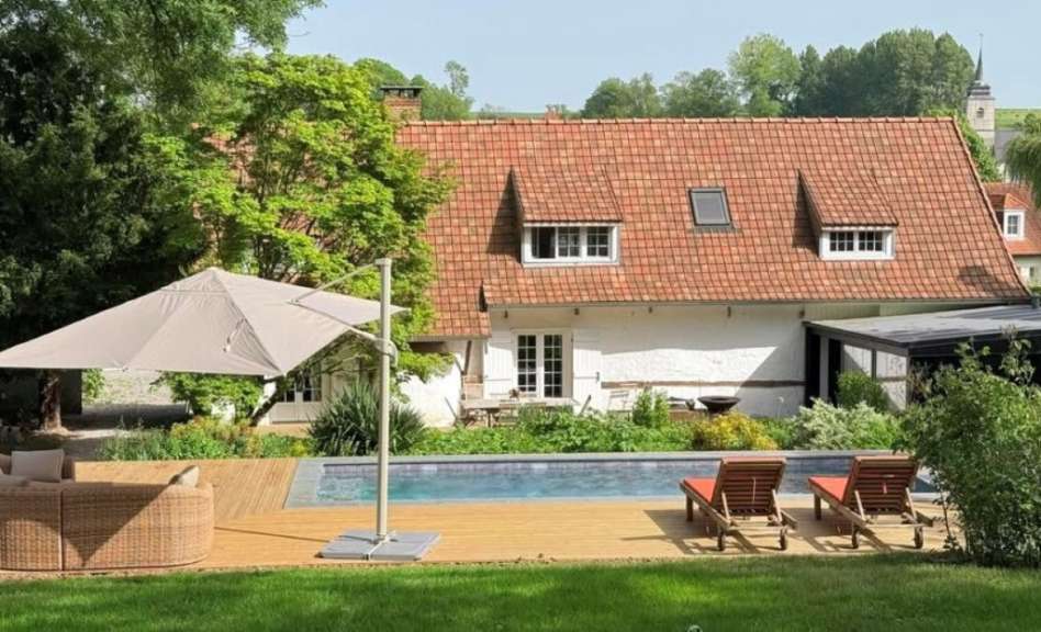 Ein weitläufiger Garten mit einem Swimmingpool und Liegestühlen vor einem weißen Haus mit rotem Ziegeldach an einem sonnigen Tag.