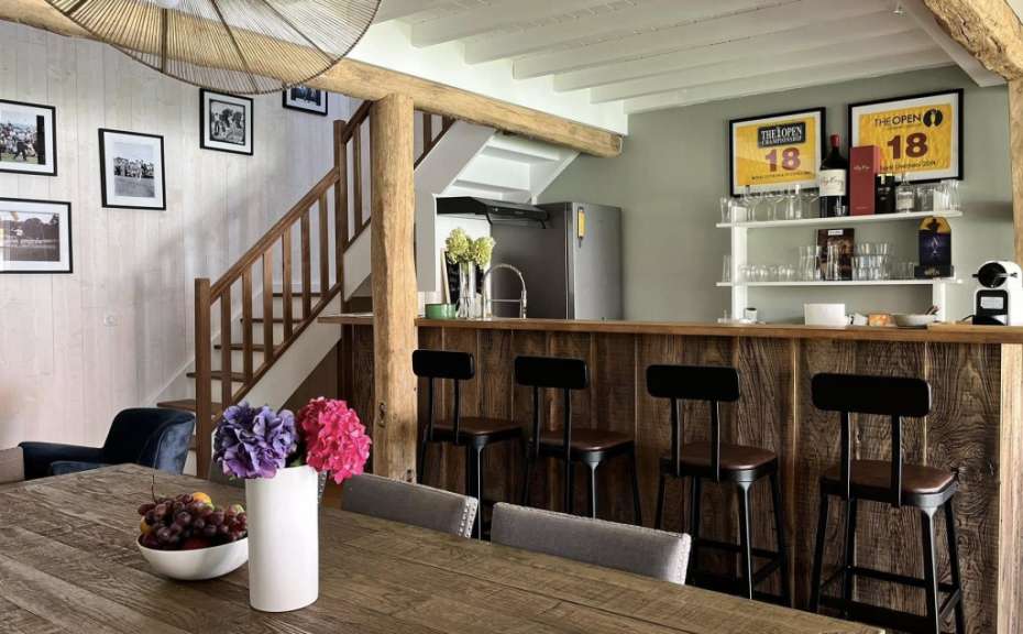 Ein rustikaler Essbereich mit einem Holztresen und Barhockern, der an eine Treppe angrenzt, ist in der Nähe eines Esstisches mit einer Obstschale und einer Vase aufgestellt.