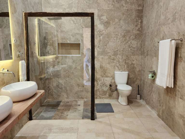 Ein geräumiges Badezimmer mit Steinzeugfliesen an den Wänden und auf dem Boden, einer bodentiefen Dusche, einem Bidet, zwei Aufsatzwaschbecken auf einer Holzplatte und einem Bidet, das von der Seite aufgenommen wurde.