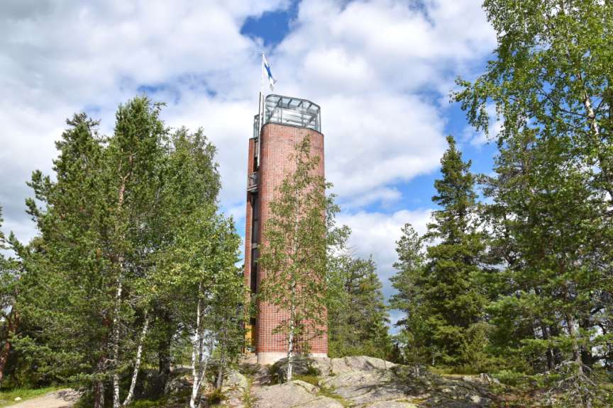 Ein roter Ziegelsteinturm mit einer Glaskuppel, auf dem eine finnische Flagge weht, steht inmitten von Bäumen auf einem felsigen Hügel unter einem bewölkten Himmel.