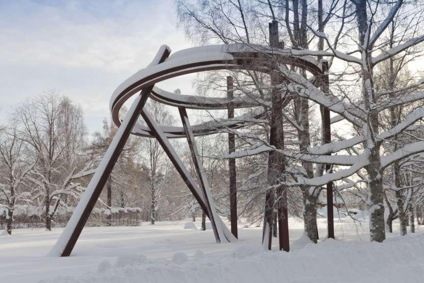Ein aufwendiges Stahlkunstwerk steht in einer verschneiten Landschaft, umgeben von Bäumen, die ebenfalls mit Schnee bedeckt sind.