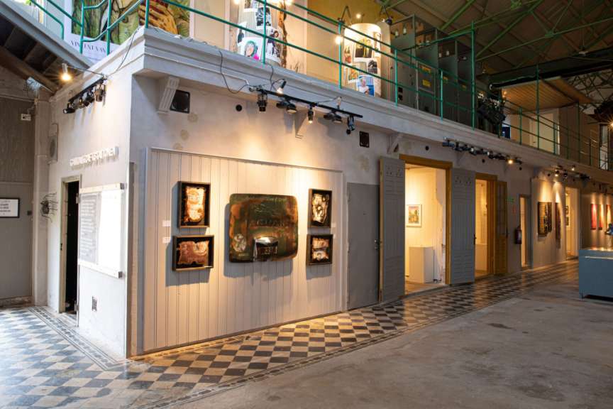 Das Bild zeigt eine Ausstellung in einer alten Kraftwerksstation mit Kunstwerken an den Wänden und Fotos im oberen Stockwerk.
