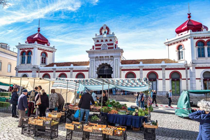 Ein belebter Marktplatz mit Obst- und Gemüseständen vor einem prächtigen Gebäude mit roten Kuppeln und Bögen.