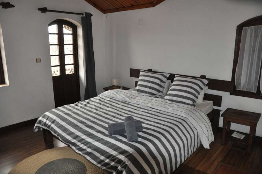 Ein großes Bett mit gestreifter Bettwäsche steht in einem Schlafzimmer mit Holzbalkendecke und einem Ausgang durch eine Tür mit Fenstern.