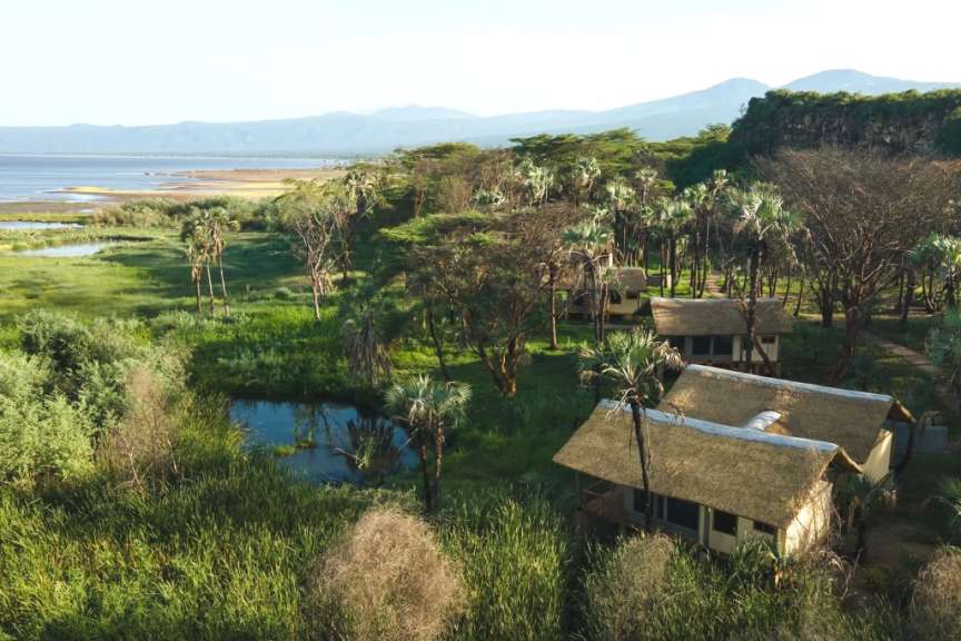 Eine Luftaufnahme zeigt strohgedeckte Bungalows inmitten üppiger grüner Vegetation, Palmen und einer weitläufigen Wasserlandschaft im Hintergrund.