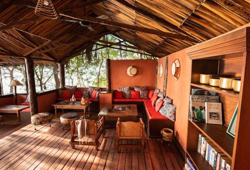Ein rustikales, offenes Wohnzimmer mit Holzböden, einem Strohdach und einer L-förmigen roten Couch ist mit Büchern und Dekorationen gefüllt.