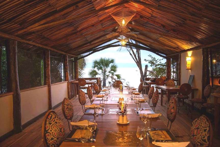 Eine lange Tafel mit Sitzgelegenheiten an beiden Seiten ist in einer Hütte mit Strohdach gedeckt, mit Blick auf das Meer und Palmen im Hintergrund.