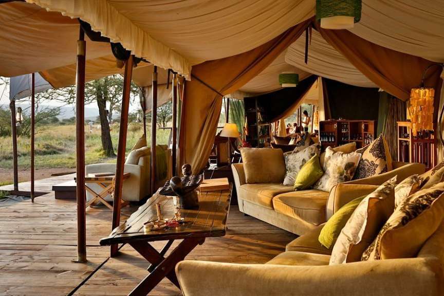 Ein luxuriöses Safari-Zelt mit bequemen Sofas und einem Holztisch ist auf einer Holzplattform im Freien aufgebaut.