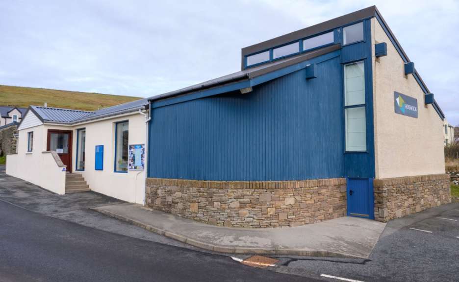 Das Bild zeigt das Hoswick Visitor Centre, ein zweigeteiltes Gebäude mit einem weißen und einem blauen Teil, das an einer abschüssigen Straße gelegen ist.