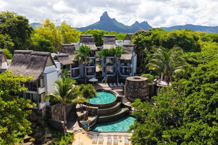 Eine Drohnenaufnahme zeigt eine Hotelanlage mit Pools und strohgedeckten Gebäuden im Vordergrund, umgeben von üppiger Vegetation und mit einem zerklüfteten Bergmassiv im Hintergrund.