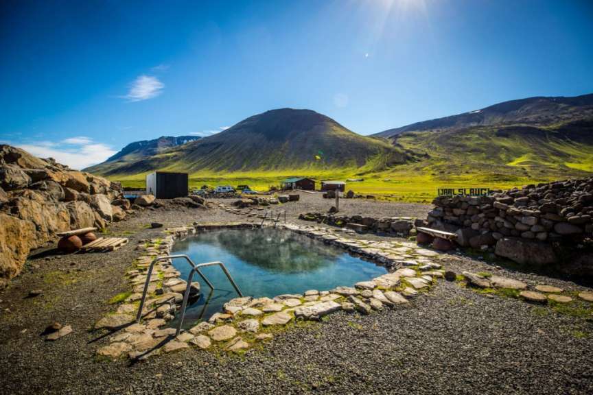 Ein geothermaler Pool mit Dampf, umgeben von Steinen und Schotter, liegt in einer grünen Landschaft unter einem sonnigen blauen Himmel.