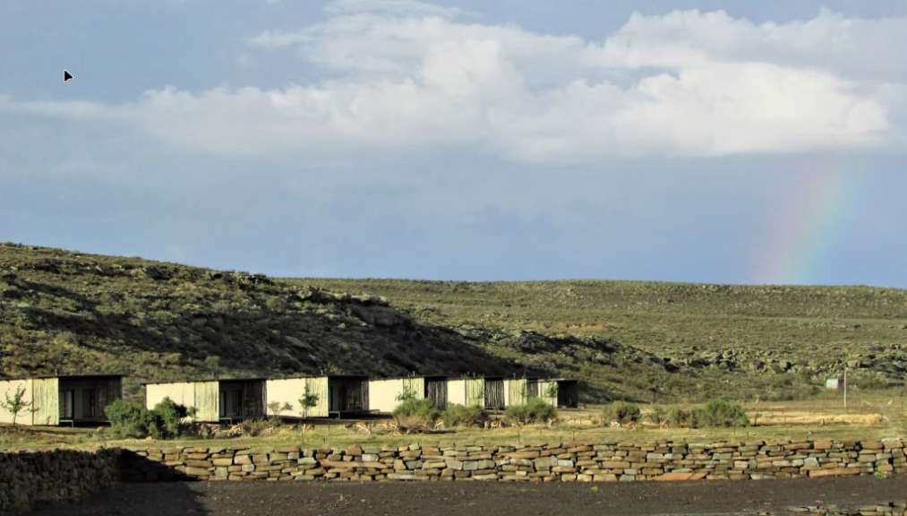 Auf diesem Bild ist eine Reihe von modernen Unterkünften in einer bergigen Landschaft zu sehen, vor der sich eine Gruppe von Antilopen auf einer grünen Wiese aufhält.