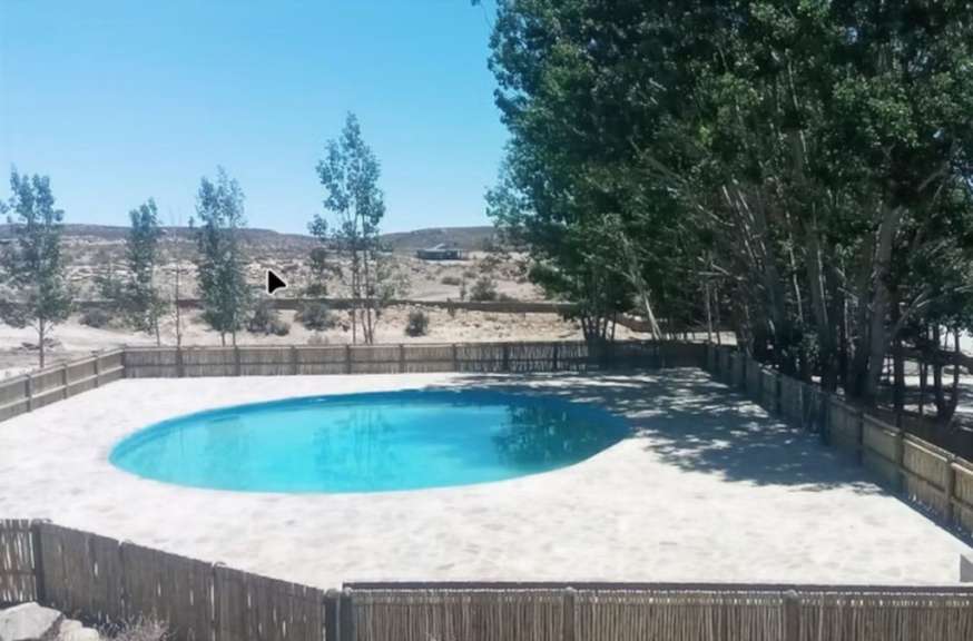 Ein ovaler Pool umgeben von einer hellen Steinplattform und einem Holzzaun, mit Bäumen und Hügeln im Hintergrund.