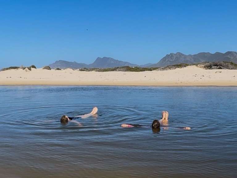 Zwei Personen treiben entspannt im Wasser, während im Hintergrund eine sandige Düne und Berge zu sehen sind.
