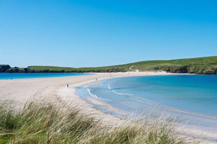 Ein breiter Sandstrand mit türkisblauem Wasser und sanften Wellen, gesäumt von grünen Hügeln unter einem strahlend blauen Himmel.