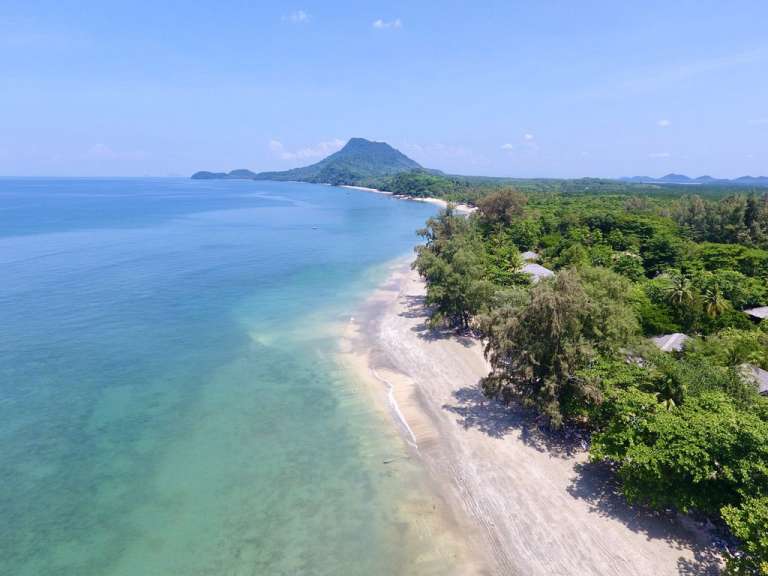 Ein Luftbild zeigt eine tropische Küstenlandschaft mit einem hellen Sandstrand, türkisfarbenem Wasser und dichtem grünen Dschungel im Hinterland, der sich bis zu einem bewaldeten Berg erstreckt.