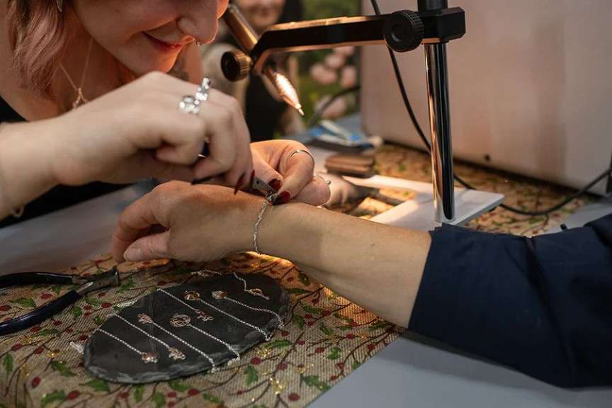 Eine Person trägt eine Brille und verwendet ein Mikroskop, um Schmuck herzustellen, während sie auf einem mit Beeren verzierten Tischtuch arbeitet.