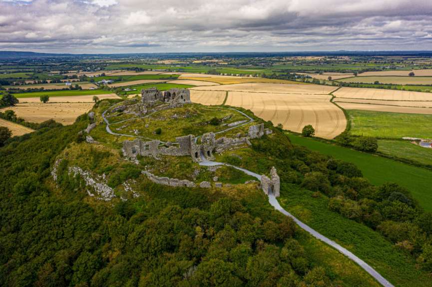 Ein verfallenes Steinschloss thront auf einem Hügel inmitten einer ländlichen Umgebung aus Feldern und grünen Hügeln unter einem bewölkten Himmel.