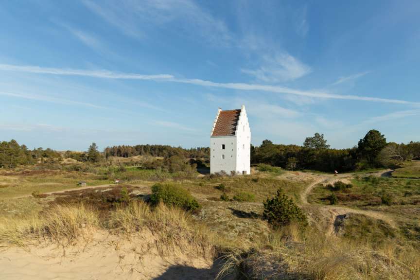 Eine weiß getünchte Kirche mit charakteristischem Treppengiebel steht auf einer bewaldeten Düne unter einem blauen Himmel.
