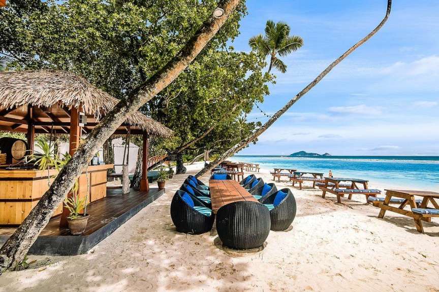 Ein Strand mit einer Bar und Tischen im Schatten von Bäumen am Meer.