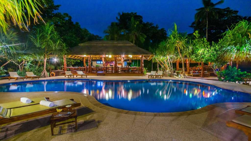 Ein beleuchteter Pool mit Liegestühlen und einem Strohdachgebäude mit Tischen und Stühlen in einem tropischen Garten bei Nacht.