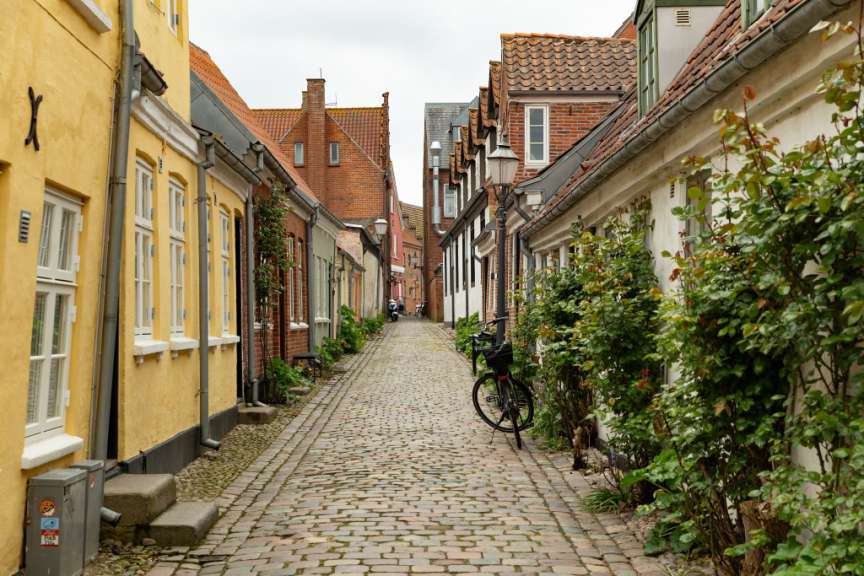 Ein Kopfsteinpflasterweg führt durch eine schmale Gasse zwischen zweistöckigen Häusern mit roten Ziegeldächern.