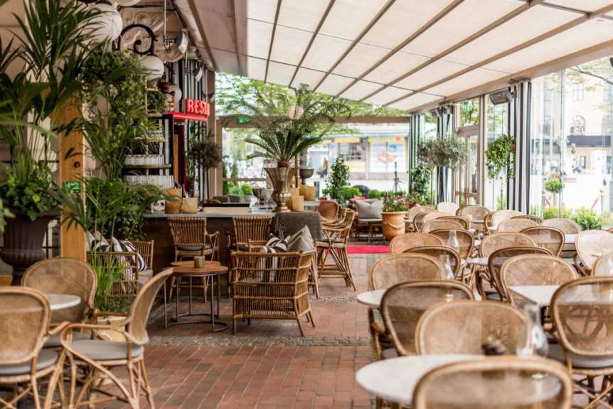 Ein offenes Restaurant mit Rattanmöbeln, Pflanzen und einer Markise ist bei Tageslicht zu sehen.