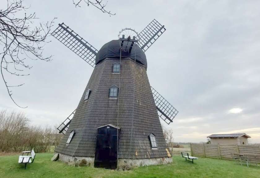 Eine Windmühle mit vier Rotorblättern steht auf einer grasbewachsenen Fläche vor einem bewölkten Himmel.