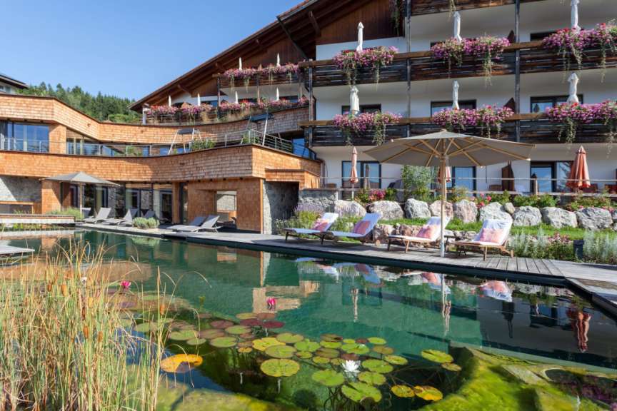 Ein mit Holzschindeln verkleidetes Gebäude mit Balkonen voller Blumen überblickt einen Teich mit Seerosen und Liegestühlen.