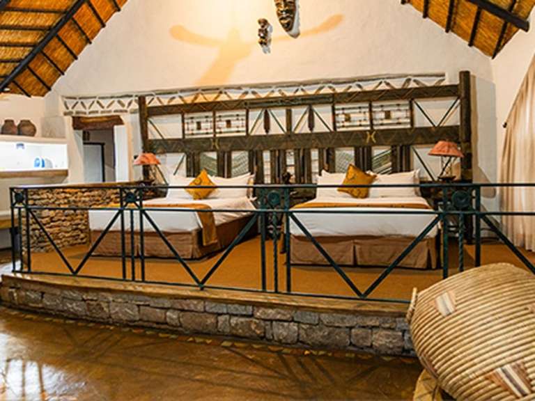 Ein Schlafzimmer mit Strohdecke und zwei Betten mit Kopfkissen, das durch einen Balkon und eine Steinmauer vom Vordergrund getrennt ist.