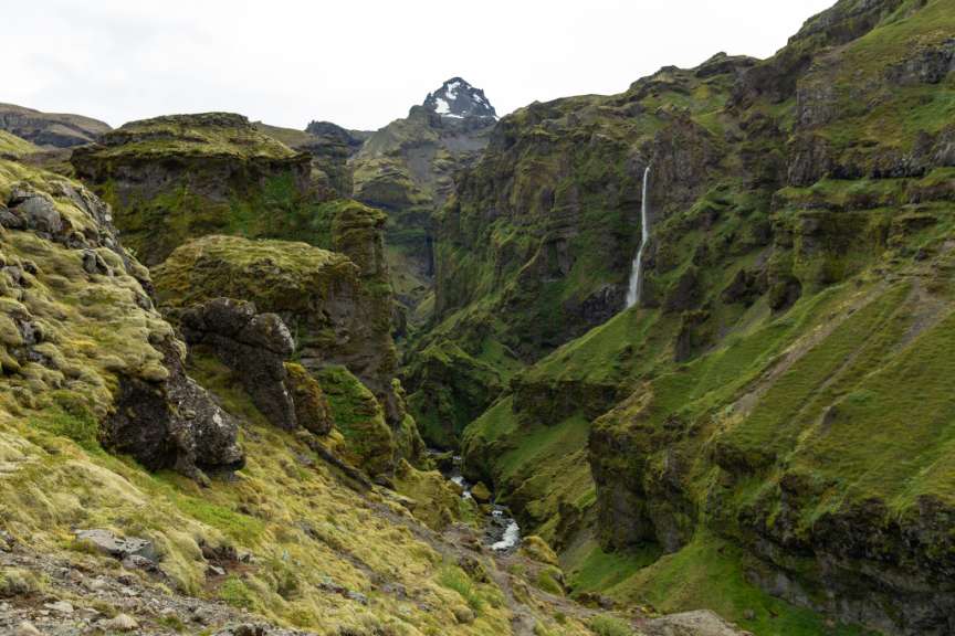 Ein langer Wasserfall stürzt in eine üppig grüne Schlucht mit bemoosten Felsen und einem Bachlauf.